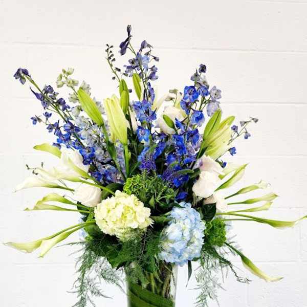 Tall blue and white flower arrangement with lilies, delphinium, and hydrangeas in a clear glass vase