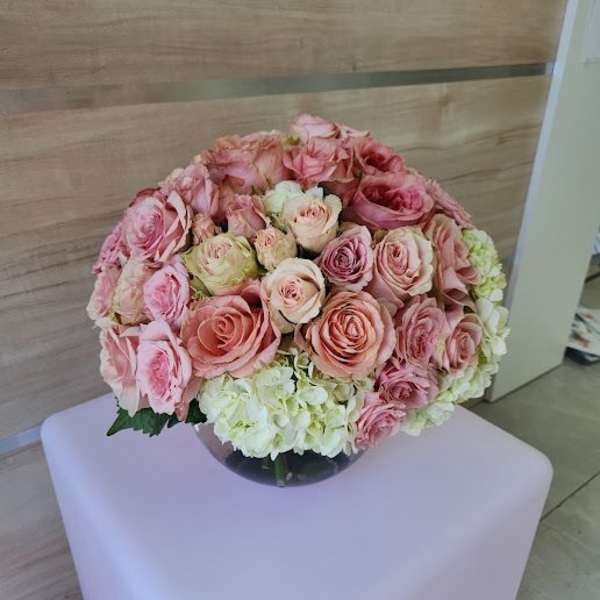 Round arrangement of pink roses and white hydrangeas in a clear glass bowl vase on a pale cube stand