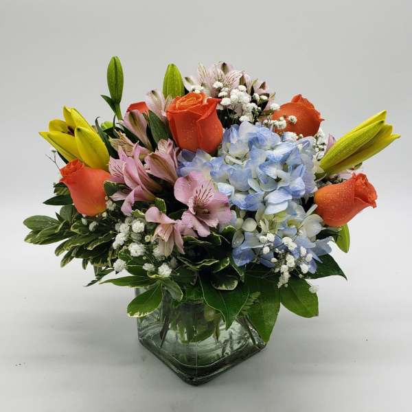 Mixed bouquet of orange roses, blue hydrangea, and yellow lilies in a glass vase