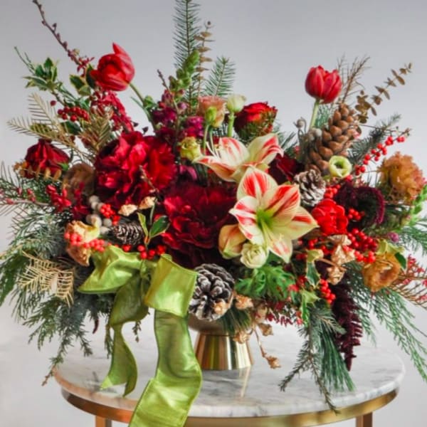 Festive red floral arrangement with amaryllis, pinecones, and a green ribbon in a gold vase