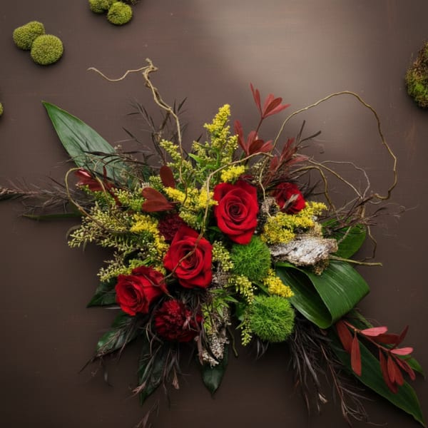 Asymmetrical arrangement of red roses with yellow accents, moss and bark on a dark brown background.