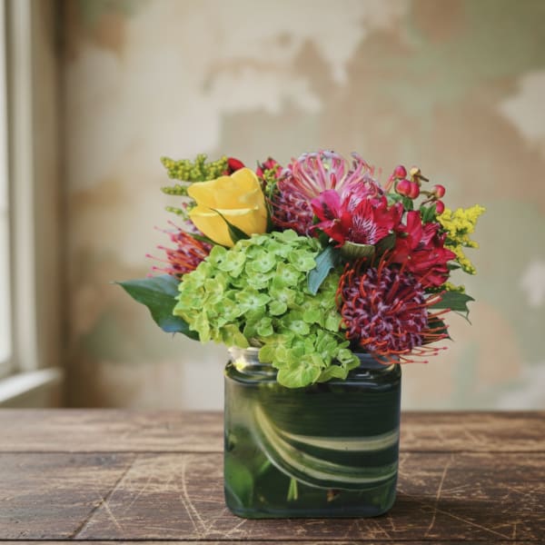 Compact arrangement of green hydrangeas, yellow calla, and red protea in a square glass vase