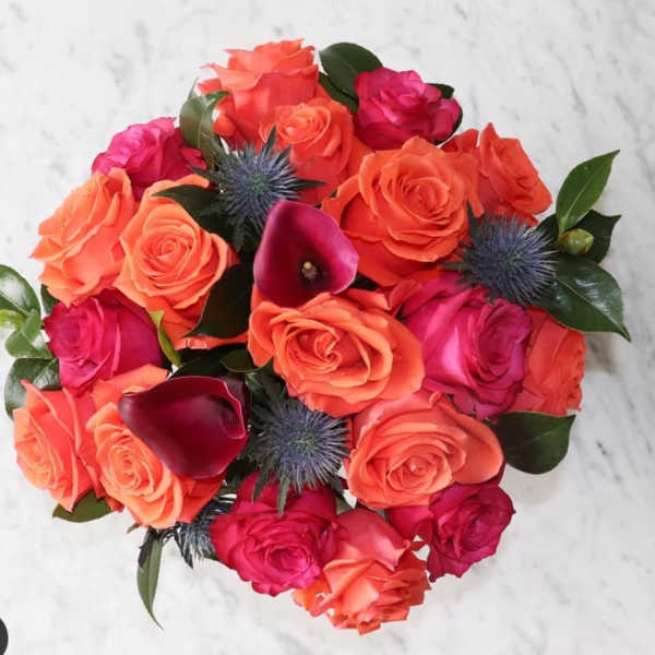 Round bouquet of bright orange and hot pink roses with dark purple calla lilies and blue thistle blooms.
