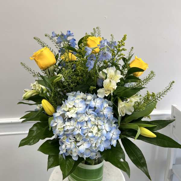 Mixed arrangement of yellow roses and tulips, blue hydrangea, and white blooms in a glass vase with green wrap