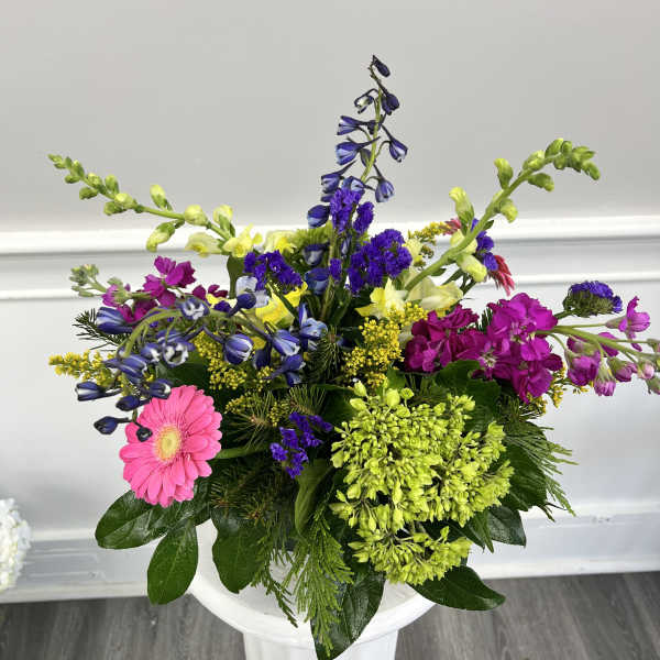 Bright mixed arrangement with pink gerbera, purple flowers, and yellow snapdragons displayed on a white pedestal.
