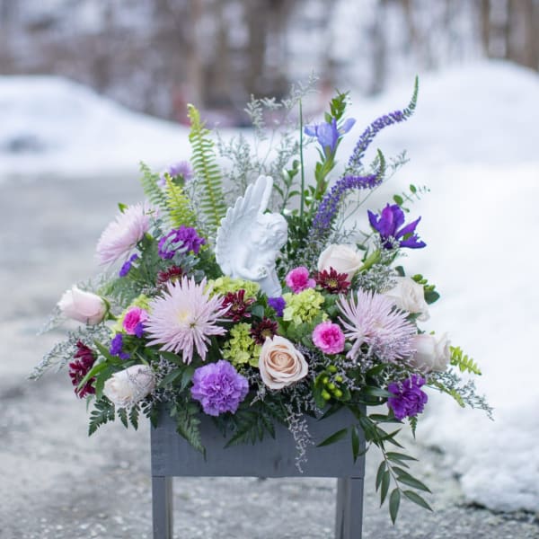 Purple and white floral arrangement in a gray container with an angel figurine