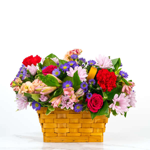 Low basket arrangement of pink daisies, red roses, red carnations, and purple asters in a woven wood basket