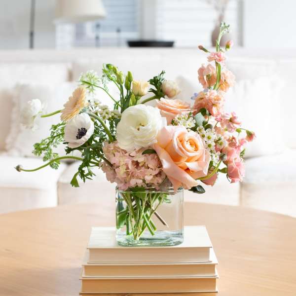 Pastel arrangement of peach roses and white blooms in a glass cube vase on books on a coffee table