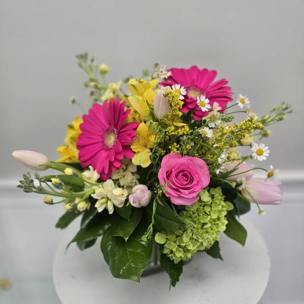 Bright mixed arrangement with hot pink gerbera daisies, pink rose, tulips, and yellow blooms in a clear glass vase