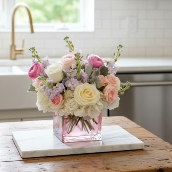 Pastel roses and ranunculus arranged in a pink glass cube vase on a wooden table.