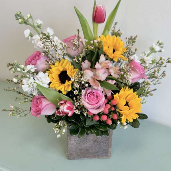 Mixed bouquet with pink roses, sunflowers, and white filler flowers in a square vase