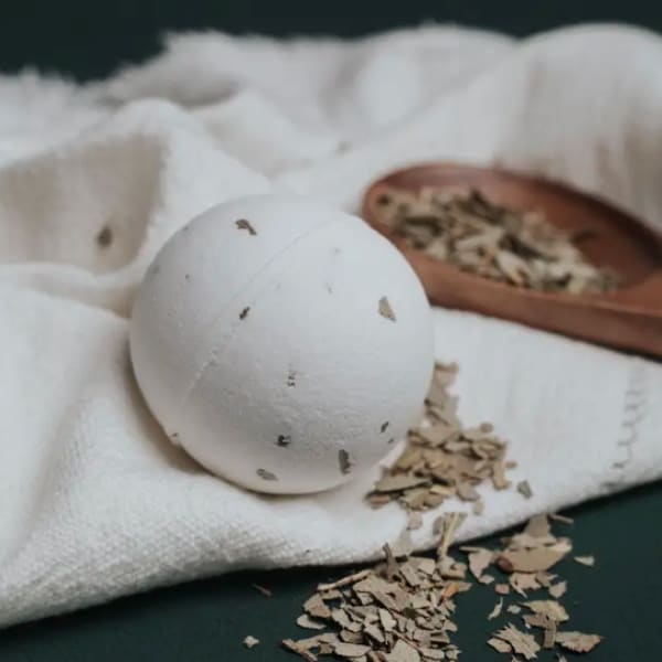 White bath bomb with herb flecks on a folded towel beside a wooden spoon of dried leaves.