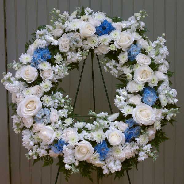 Large standing wreath of white roses and mixed white flowers with blue accents on a metal easel.