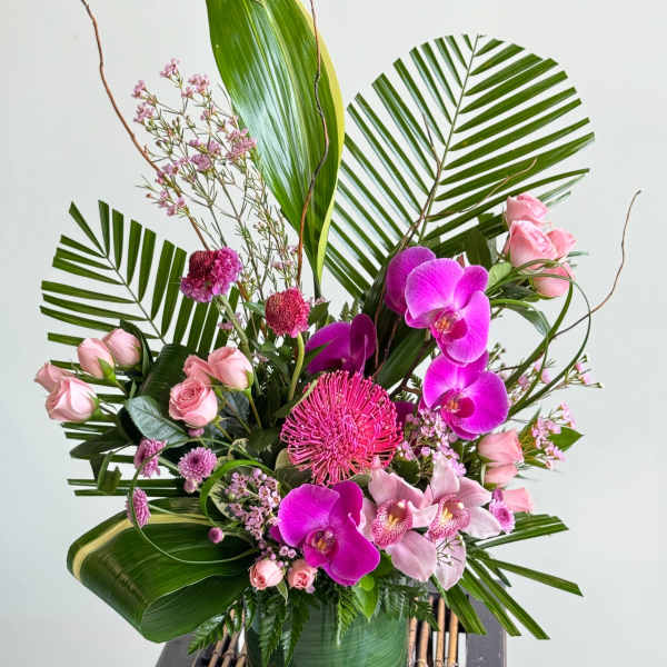 Pink orchids and roses arranged in a glass vase with large tropical leaves