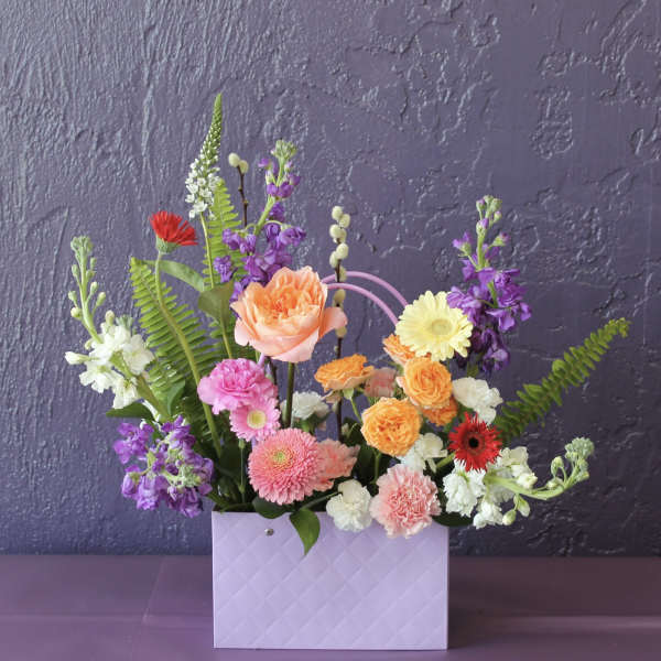 Colorful mixed flower arrangement in a lavender gift bag-style container against a purple wall