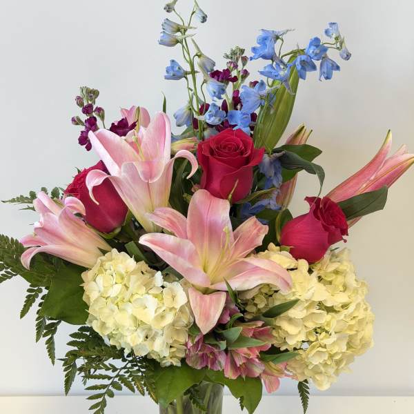 Bouquet of pink lilies, red roses, blue delphinium, and pale hydrangeas in a glass vase