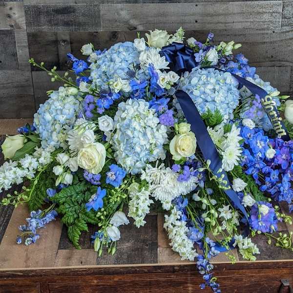 Low blue and white casket spray with hydrangeas, roses, and delphinium accented by navy ribbon.