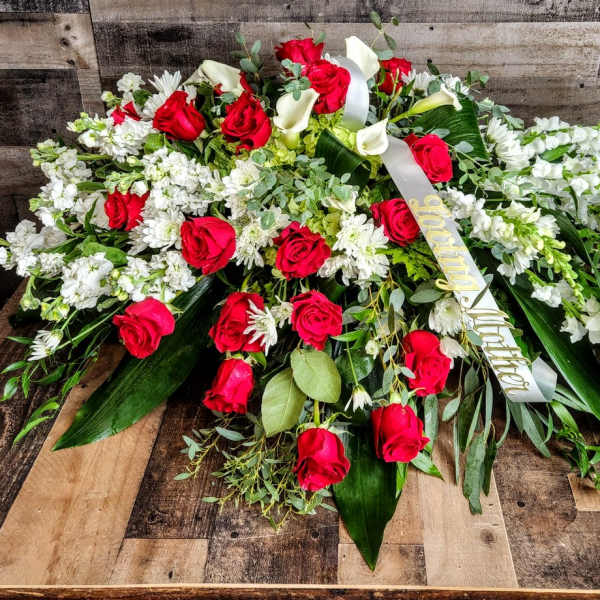 Large red and white casket spray with roses, lilies, and ribbon on a wooden surface