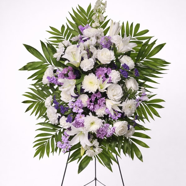 Standing spray of white lilies, roses, and purple mums with large palm leaves on a wire easel.