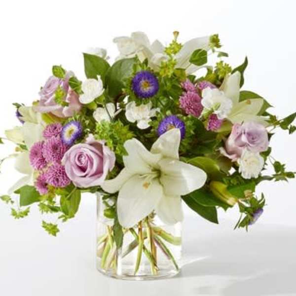 Mixed arrangement of white lilies, lavender roses, and purple asters in a clear glass vase