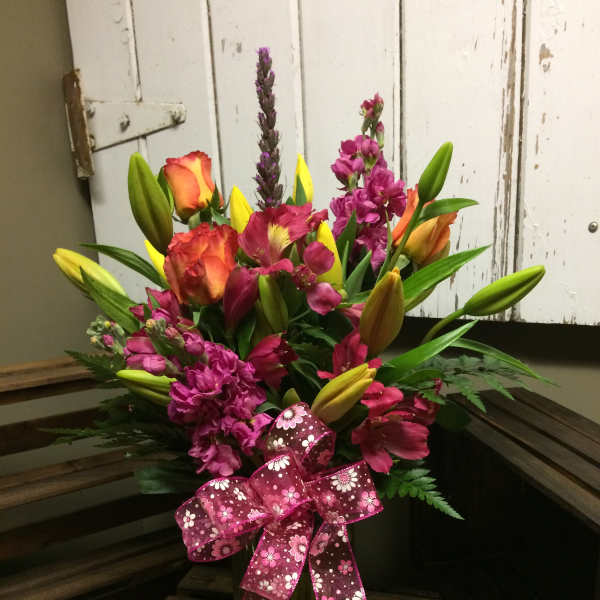Mixed bouquet of lilies, roses, and purple blooms in a glass vase with a pink ribbon