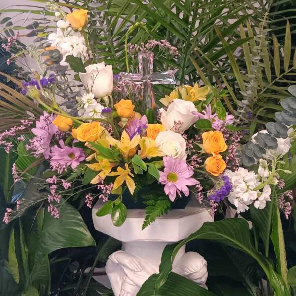 Arrangement of yellow and white flowers with a glass cross, set among tall palm greenery on a white pedestal.