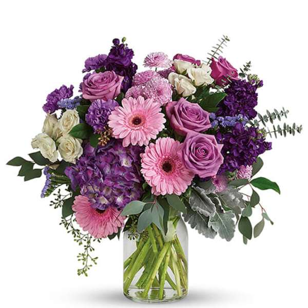 Mixed bouquet of pink gerbera daisies, lavender roses, and purple blooms in a clear glass vase