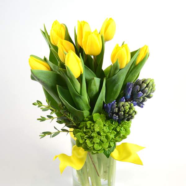 Yellow tulips arranged in a clear glass vase with green and purple flowers
