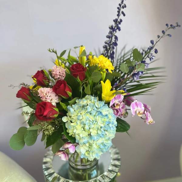 Mixed bouquet with red roses, blue hydrangea, yellow blooms, and orchids in a glass vase