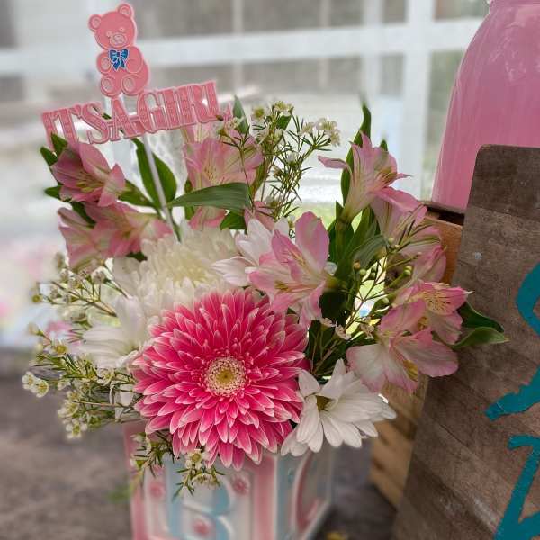 Pink and white baby-themed flower arrangement in a pastel block vase with It's a Girl pick