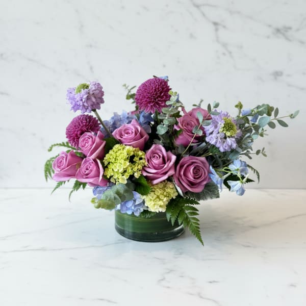 Low arrangement of mauve roses with purple mums and green and blue hydrangea in a glass vase