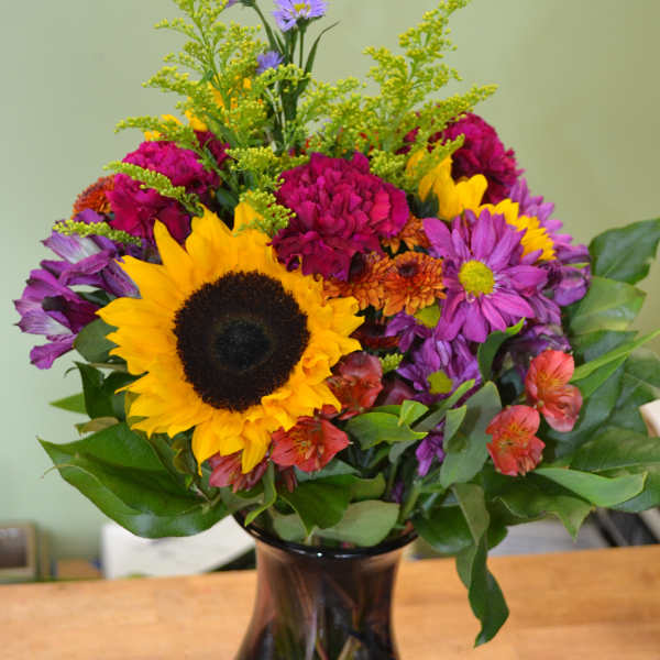 Mixed bouquet with a sunflower and purple, pink, and orange flowers in a dark glass vase