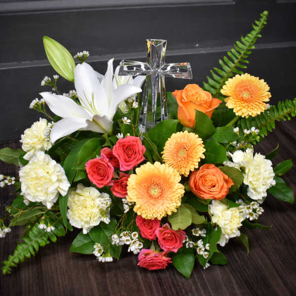 Low arrangement of white lily, peach gerberas, orange and pink roses with a clear glass cross in the center.