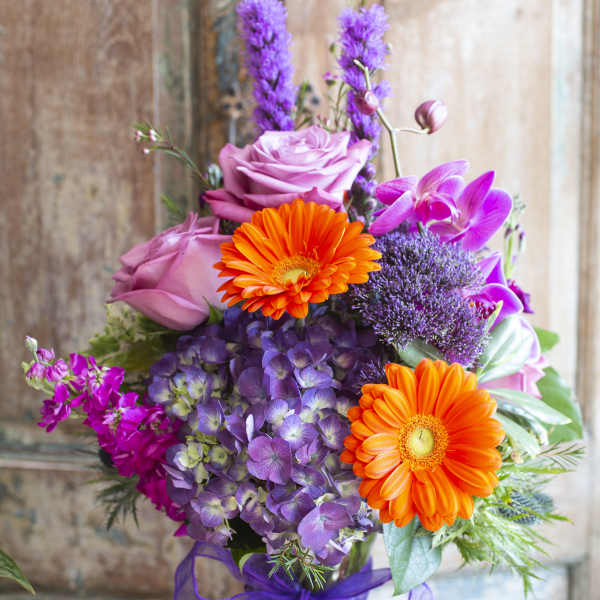Bouquet of pink roses, orange gerberas, and purple flowers in a glass vase