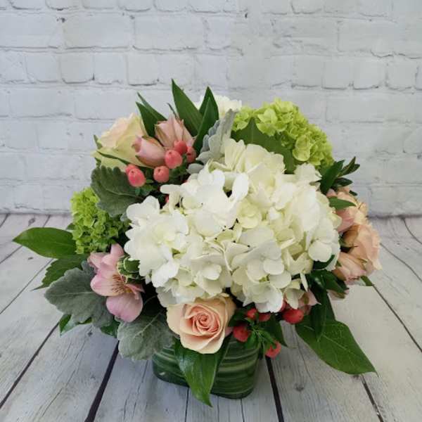 Low arrangement of white hydrangeas with peach roses and pink accents in a green glass cube vase