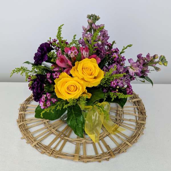 Compact arrangement of yellow roses and purple mixed flowers in a glass vase with a yellow bow