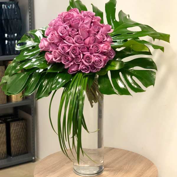 Tall glass vase with a dome of mauve roses framed by large glossy monstera leaves and trailing blades of grass