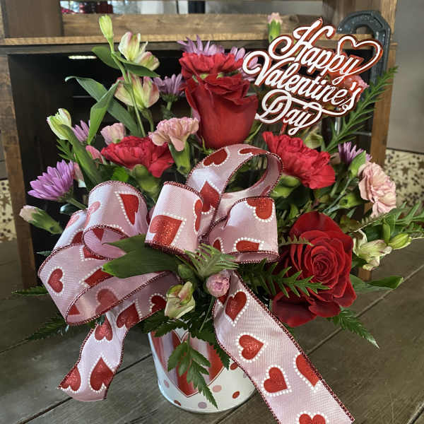 Valentine bouquet with red roses, pink carnations, and purple daisies in a heart-print tin