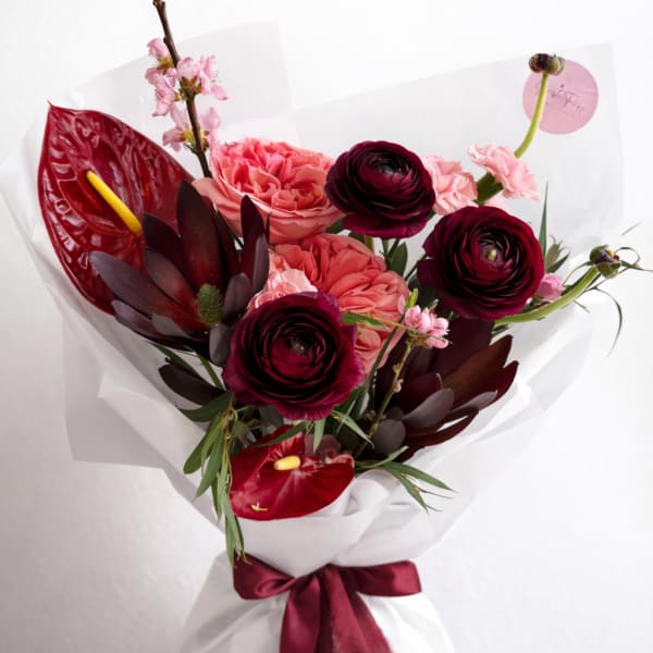 Hand-tied bouquet of dark red ranunculus, pink roses, and tropical blooms wrapped in white paper with burgundy ribbon