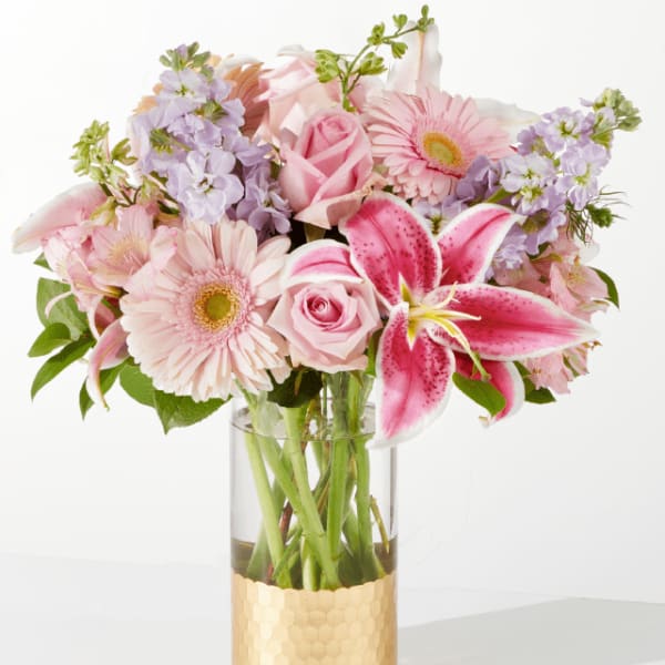 Pink lilies, roses, and gerbera daisies with lavender stock in a glass vase with a gold base.