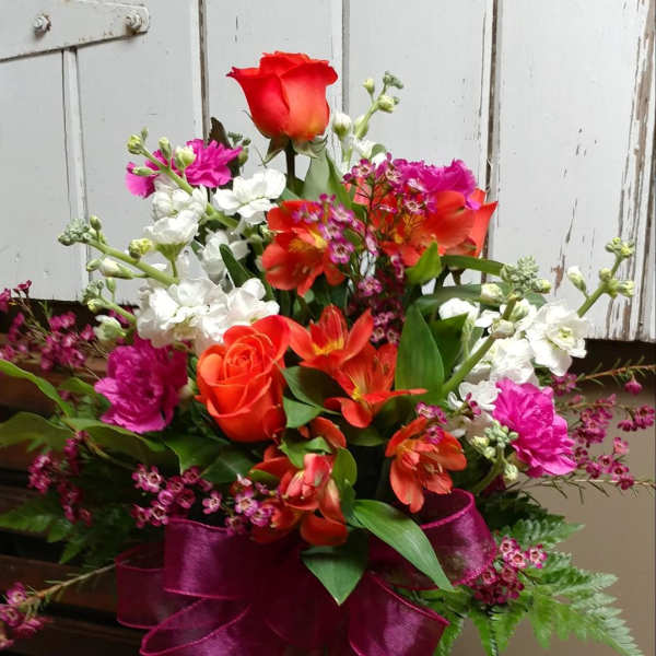 Mixed bouquet of red, orange, white, and pink flowers in a glass vase with a magenta ribbon
