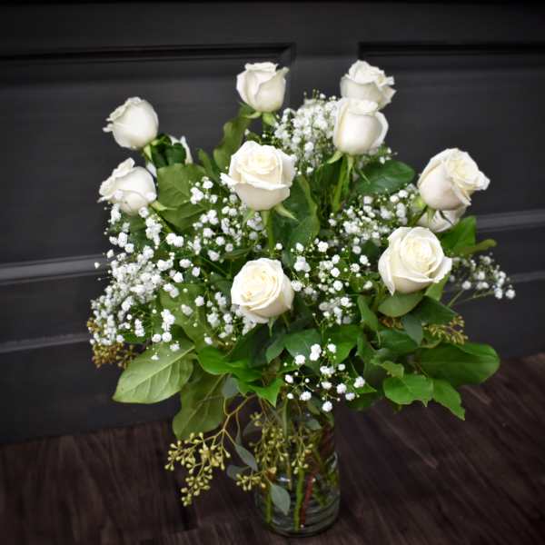 Tall arrangement of white roses and small white filler flowers in a clear glass vase.