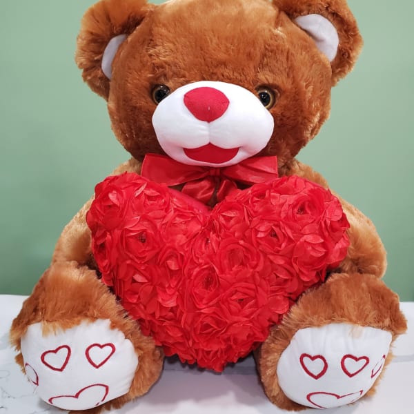 Brown teddy bear holding a red heart-shaped floral decoration
