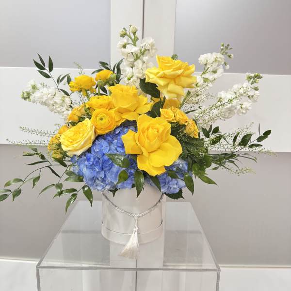 Yellow roses and blue hydrangeas in a white hatbox with white flowers.