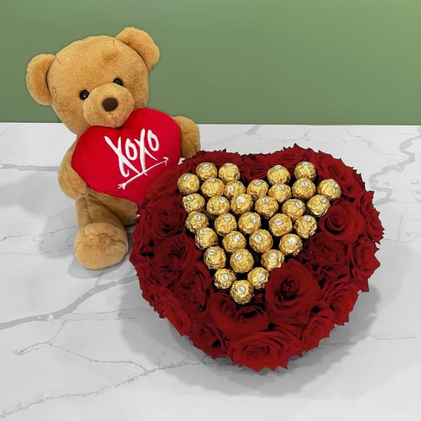 Teddy bear holding a red heart beside a heart-shaped rose and chocolate arrangement