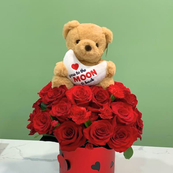 Teddy bear holding a heart pillow above red roses in a heart-decorated box