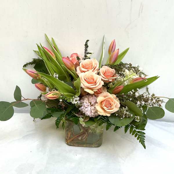 Peach roses and pink tulips arranged in a low clear glass cube vase with white filler flowers.