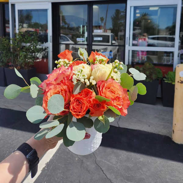 Compact arrangement of orange and cream roses in a small white vase held outdoors
