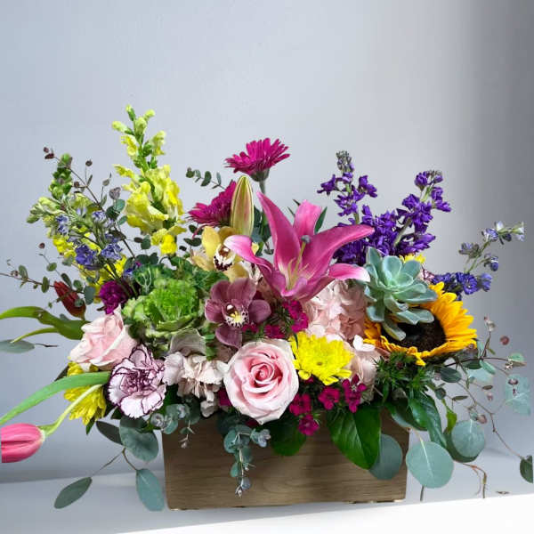 Colorful mixed flower arrangement with lilies, roses, sunflowers, succulents, and other blooms in a wooden box.