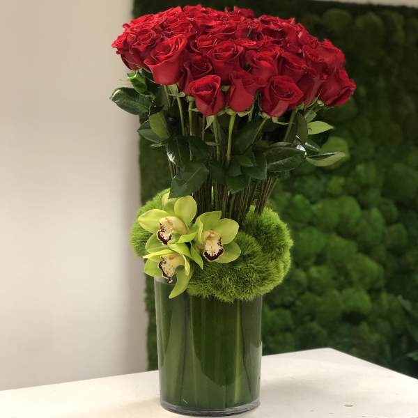 Tall arrangement of red roses and green cymbidium orchids in a clear glass vase wrapped with green